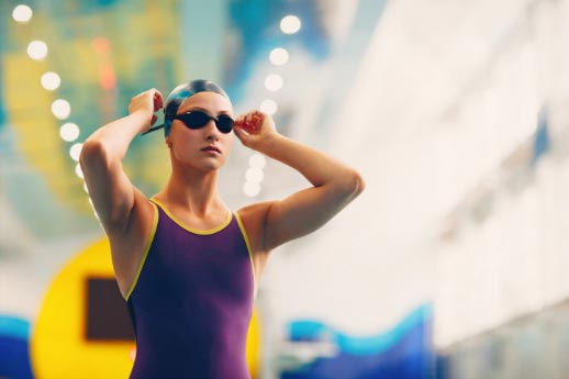 A woman swimmer with a cap and her hands on her goggles at the edge of the pool.